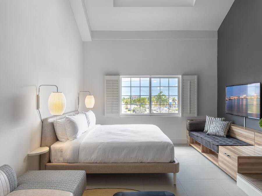 Un dormitorio moderno con cama, mesitas de noche, televisión y ventana con vista a la ciudad.