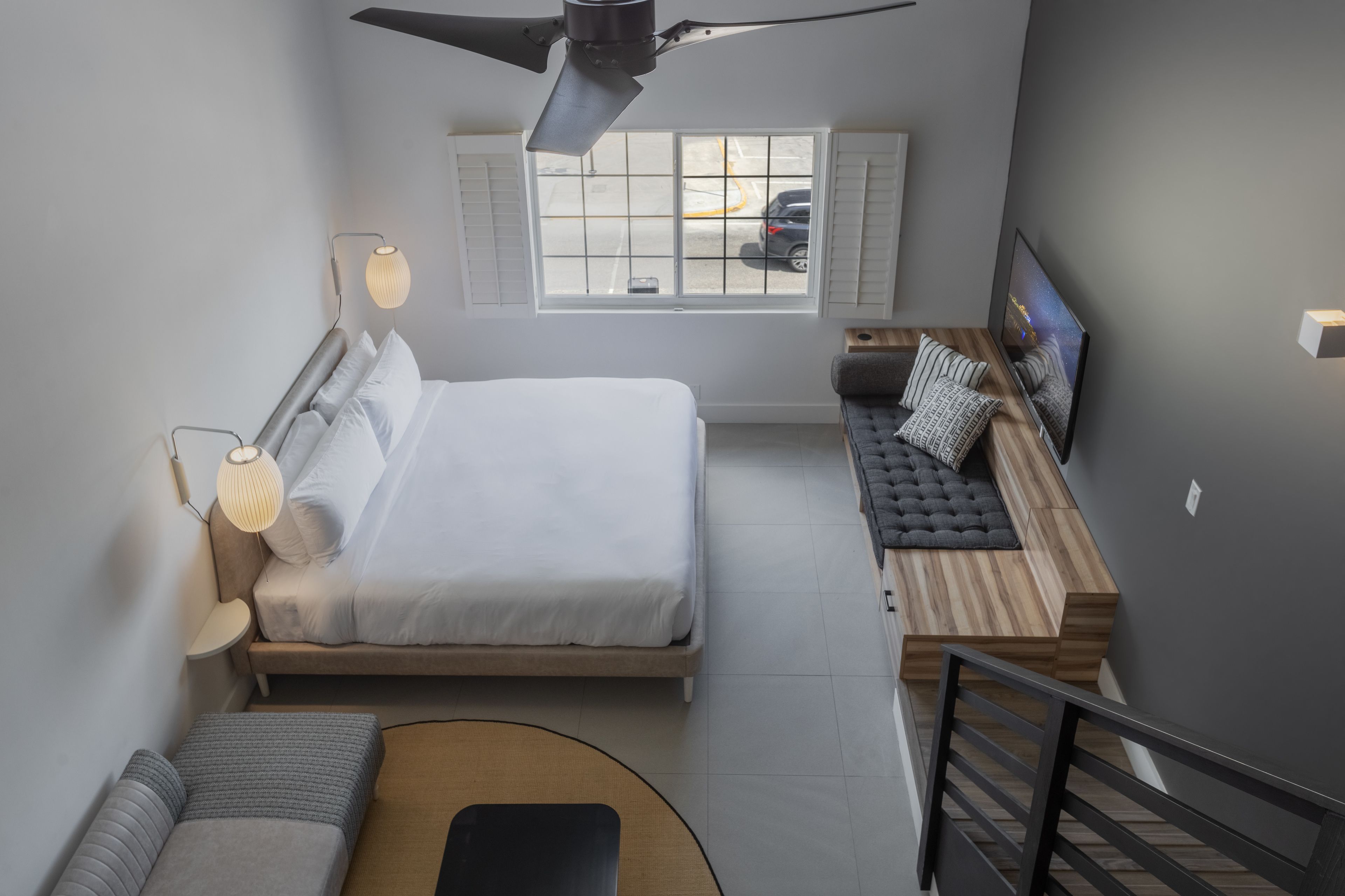 Un dormitorio moderno con cama, zona de estar con televisor, ventana con persianas y ventilador de techo.