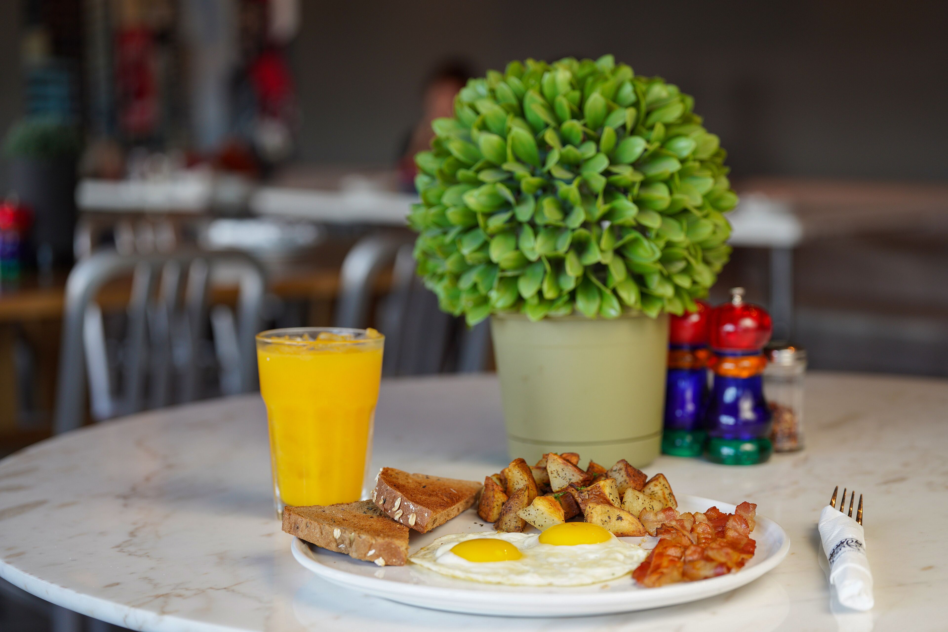 A breakfast plate with eggs, toast, and potatoes, a glass of orange juice, and a small potted plant on a marble table.