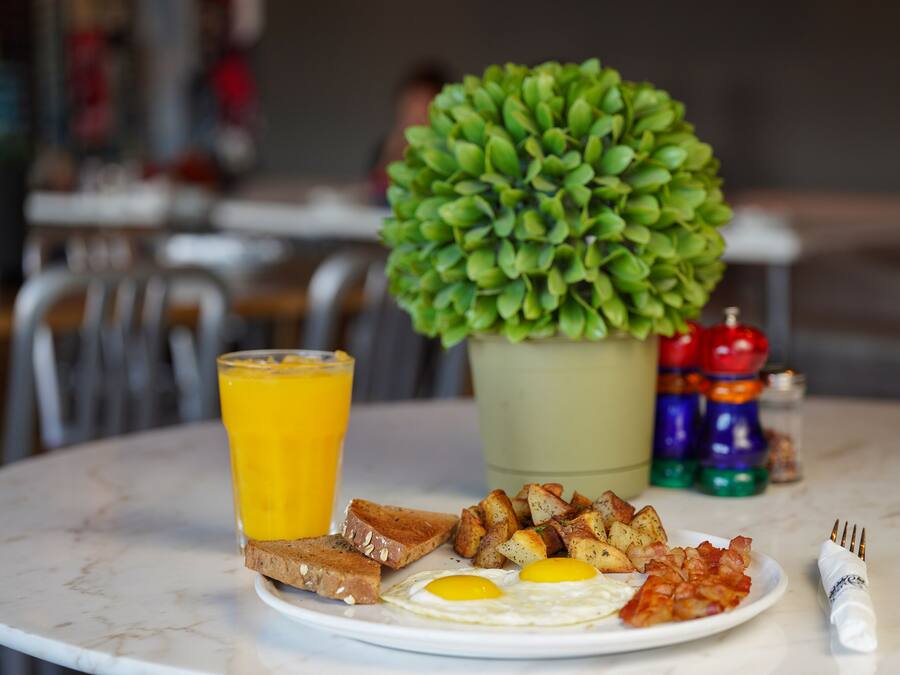 Un plato de desayuno con huevos, tostadas y papas, un vaso de jugo de naranja y una pequeña planta en maceta sobre una mesa de mármol.