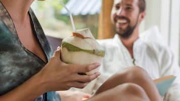 Una persona sostiene un coco con una pajita, otra persona está sentada al fondo, y una ventana muestra una vista de follaje.