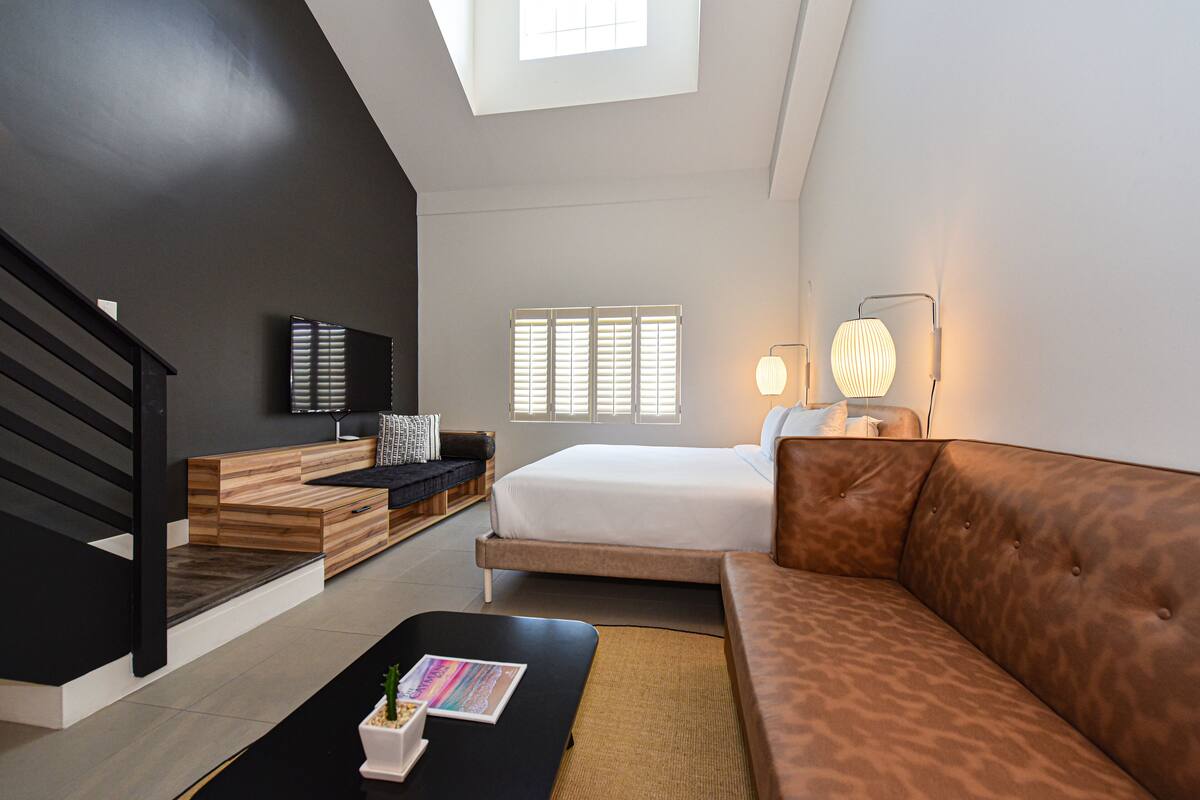 Ein modernes Hotelzimmer mit Loft-Charakter, einem Bett, einer Couch, einem Couchtisch und einem Fernseher.