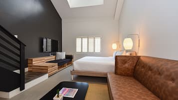 A modern hotel room with a loft-style ceiling, a bed, a sofa, a coffee table, and a TV.