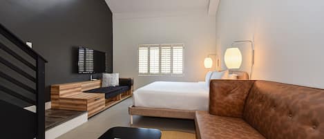 A modern hotel room with a loft-style ceiling, a bed, a sofa, a coffee table, and a TV.