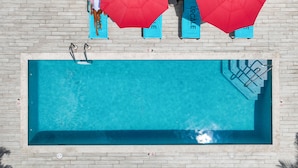 Una piscina rectangular con una terraza de baldosas, rodeada de palmeras y sombrillas rojas.