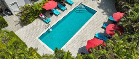 Un área de piscina en la azotea con una piscina rectangular, rodeada de sillones azules y sombrillas rojas, con palmeras y un edificio al fondo.