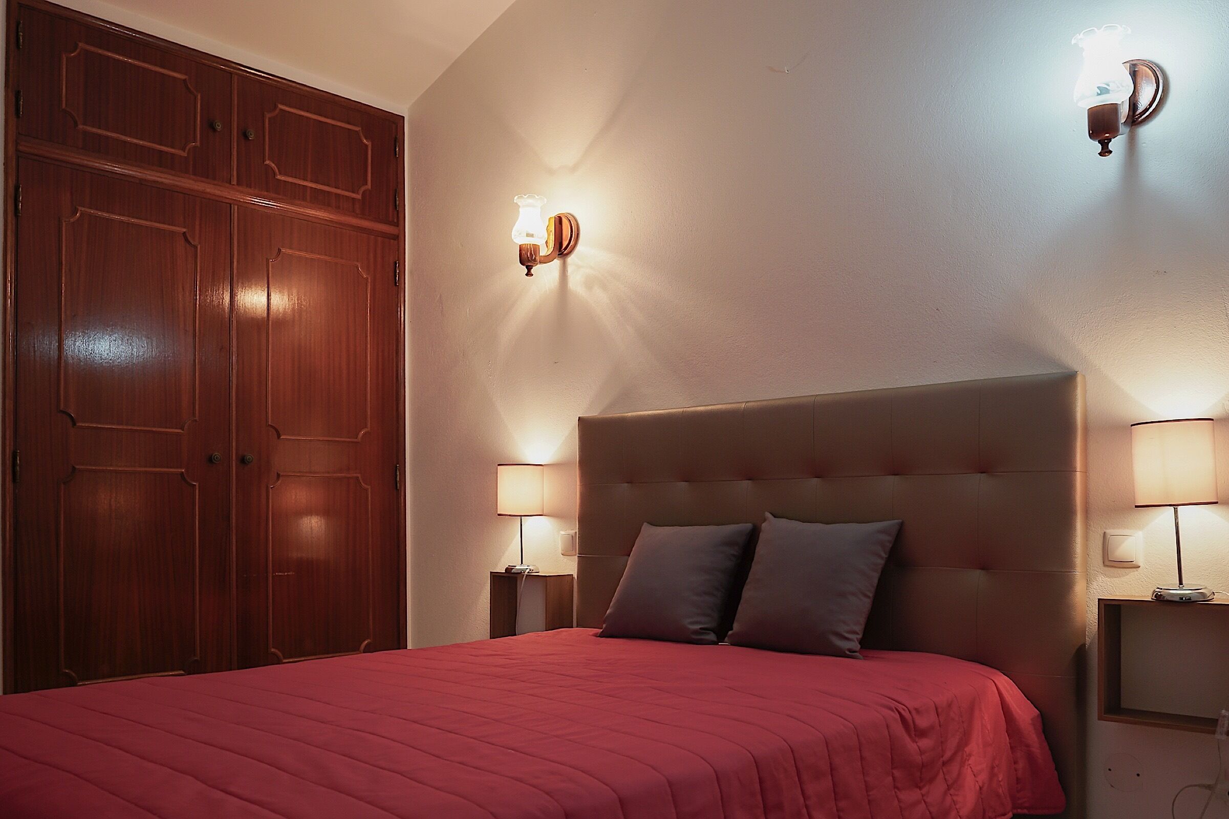 A bedroom with a large bed, wooden wardrobe, two bedside lamps, and a wall-mounted light fixture.