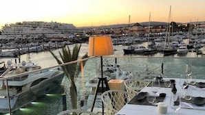 Ein Yachthafen mit vertäuten Booten, ein für das Essen gedeckter Tisch mit Gläsern und Besteck, und eine Lampe auf dem Tisch.