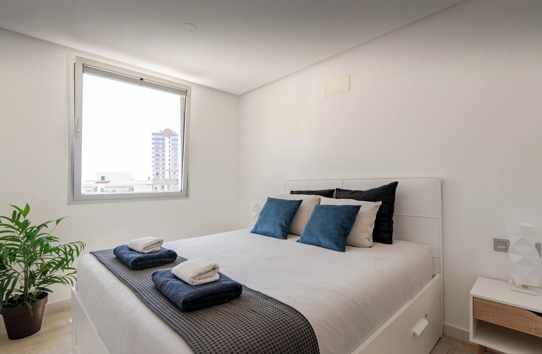 A modern bedroom with a large bed, a potted plant, and a view of a cityscape through a window.