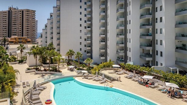 Un hôtel moderne doté d’une piscine centrale, entourée d’espaces de détente et de palmiers.