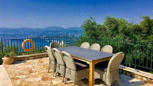 An outdoor dining area with a table and wicker chairs, overlooking a scenic view of the sea and mountains. - Prestige Villas |  SPARTILAS | Secluded Villa 15-20 mins walk to Taverna (Spartilas)