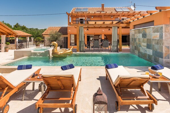 A pool area with lounge chairs, a floating swan, and a building with a tiled roof. - Holiday Home La Verna with Pool (Skradin)