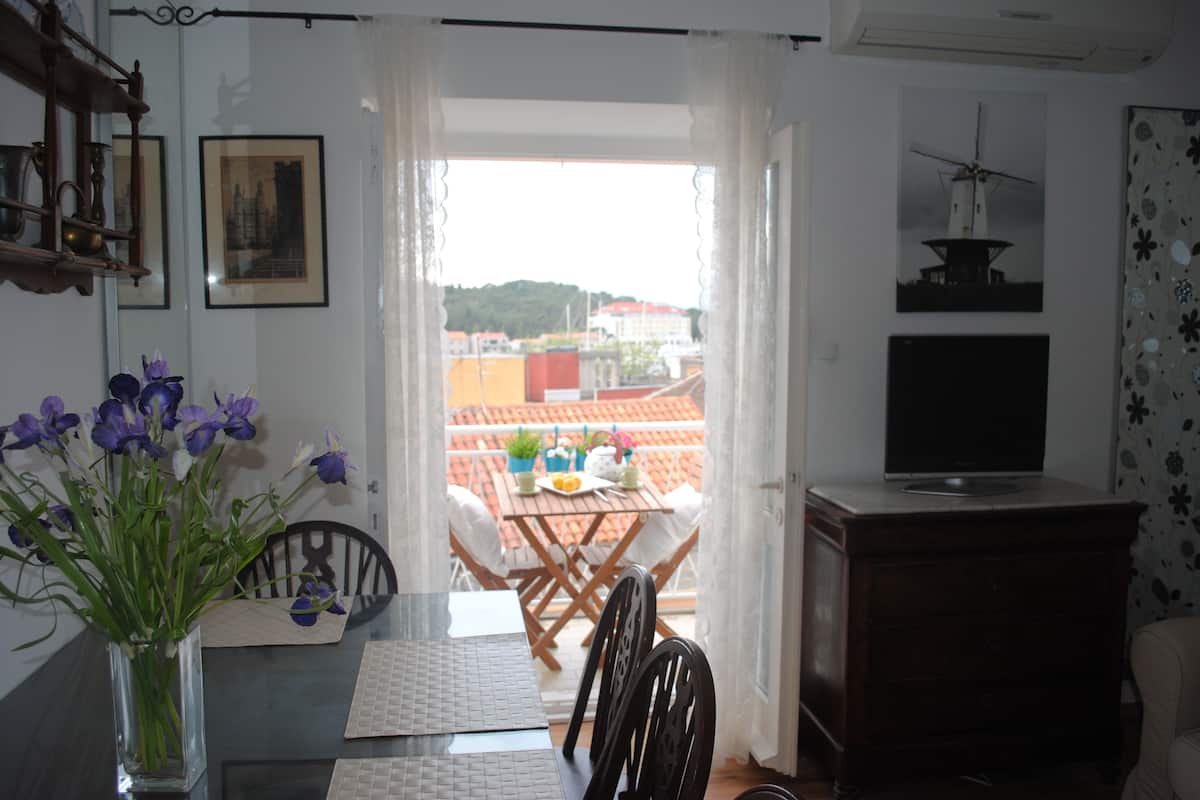 Un coin repas comprenant une table, des chaises, un vase de fleurs, un téléviseur et une vue sur les bâtiments à travers une fenêtre.