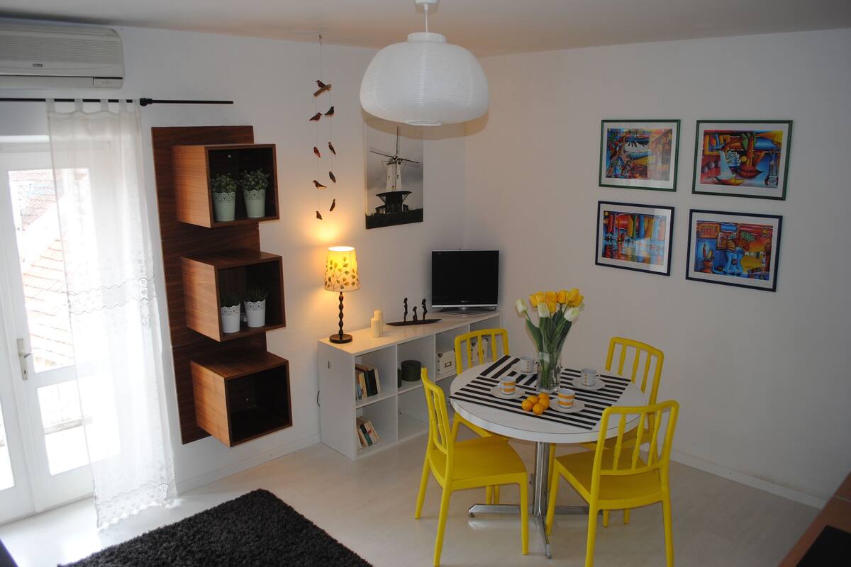 Un coin repas avec une table ronde, des chaises jaunes et un luminaire suspendu blanc.