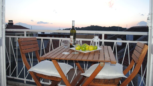 A wooden table with a bottle, glasses, and green apples on a balcony with white railings and wooden chairs. - Apartments Optimist (Makarska)