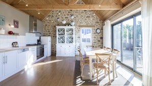 Una cocina con vigas de madera, pared de piedra, zona de comedor con mesa y sillas, y vistas a través de amplias ventanas.