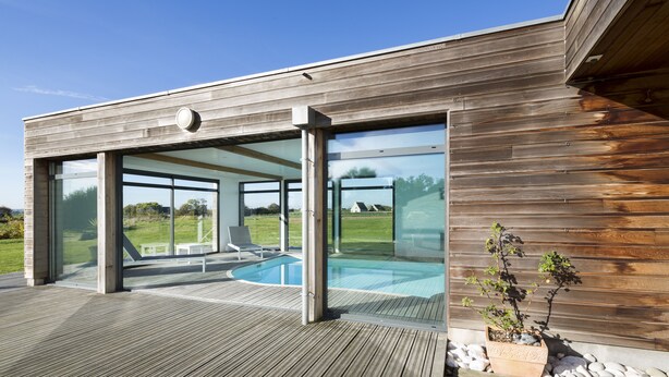 Una casa moderna con fachada de madera, grandes puertas de cristal que dan a un área de piscina al aire libre y una planta en maceta en la terraza.
