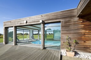 A modern house with a wooden exterior, large glass doors leading to an outdoor swimming pool area, and a potted plant on the deck. - Character house with heated indoor pool 800 m from the sea (Plouescat)