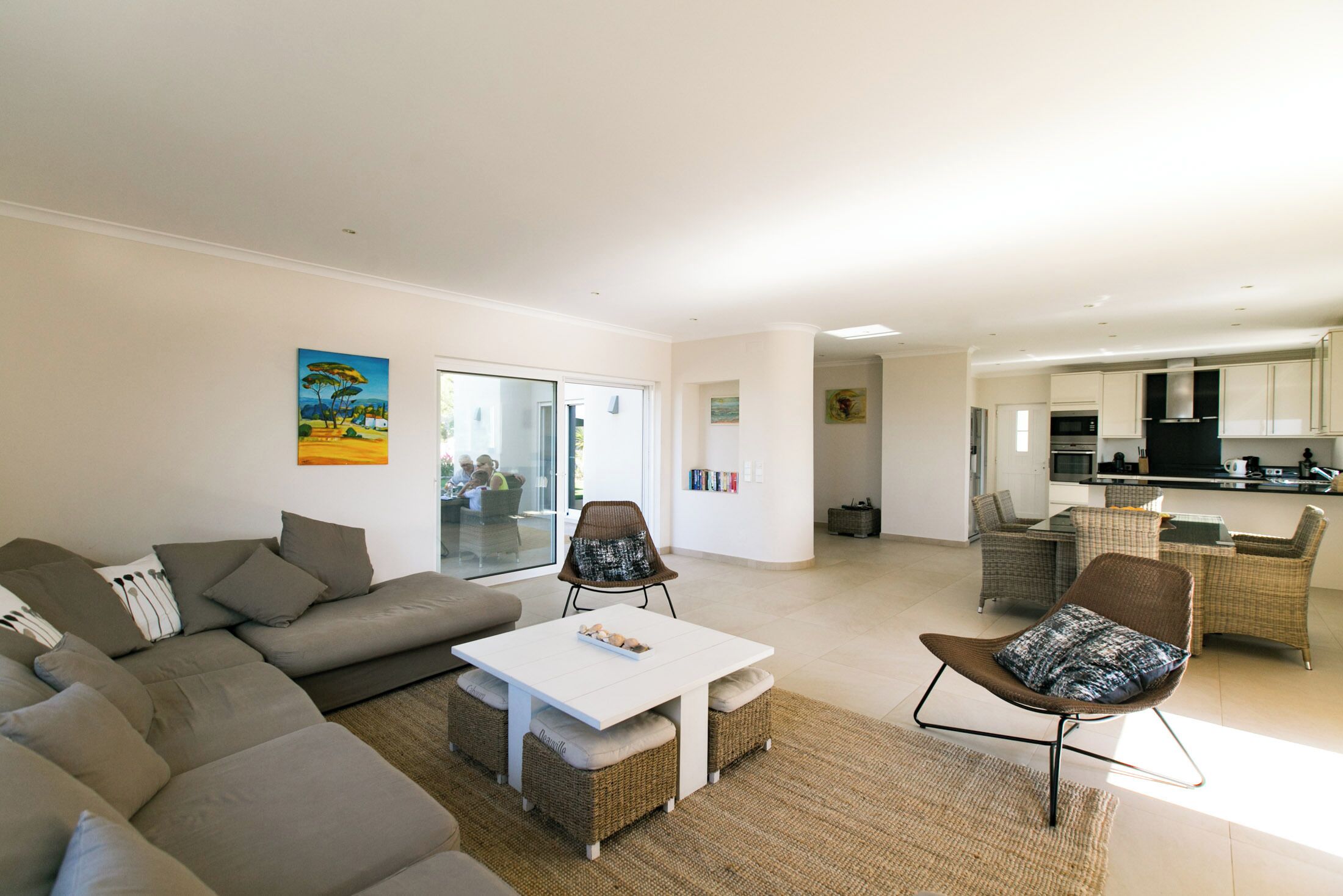 A modern living room with a sectional sofa, a white coffee table, wicker chairs, and a kitchen in the background.