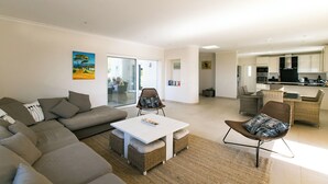 Ein modernes Wohnzimmer mit einer Eckcouch, einem weißen Couchtisch, Korbstühlen und einer Küche im Hintergrund.