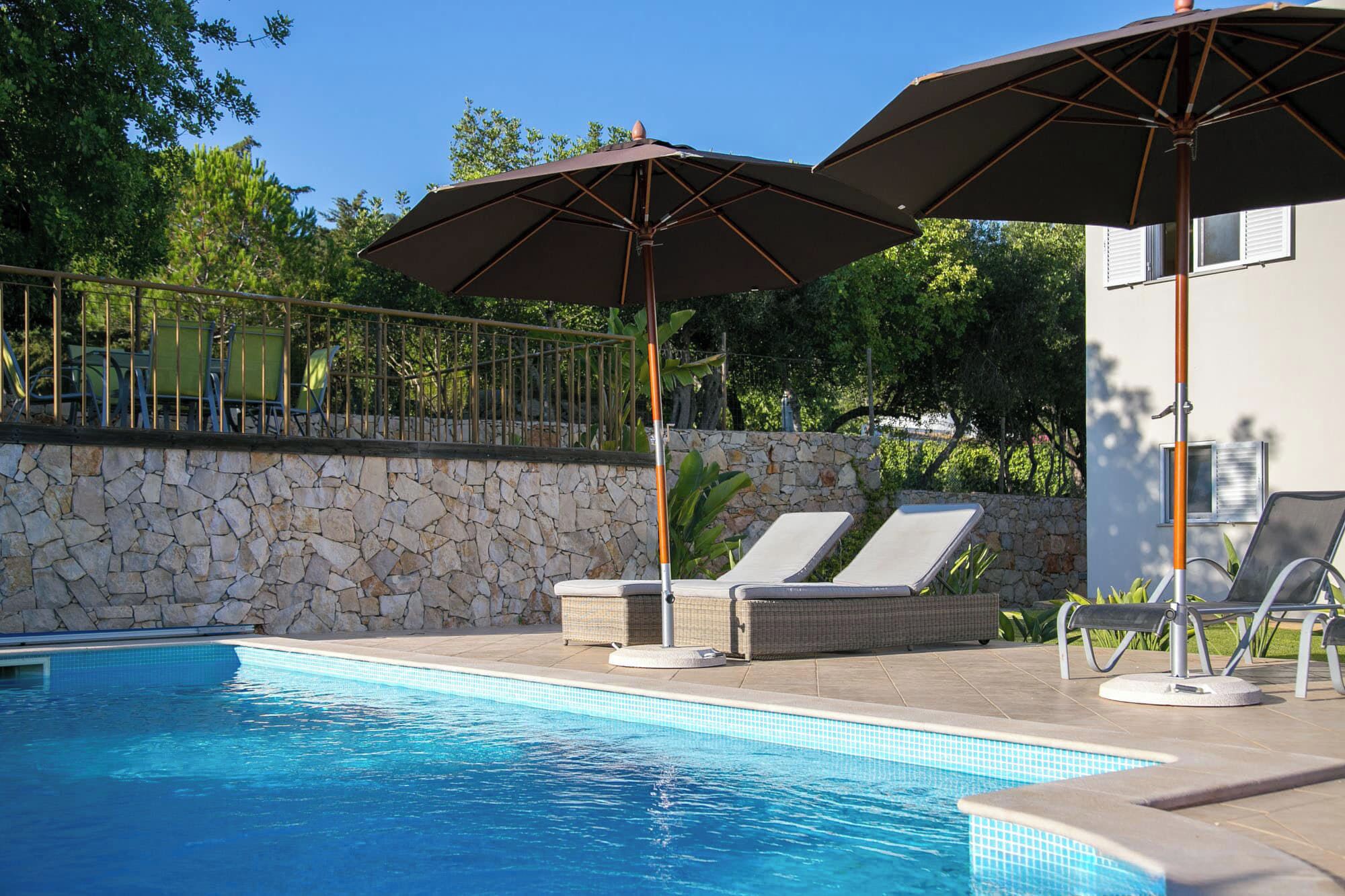 Piscina con sillones de descanso y sombrillas, rodeada de un muro de piedra y follaje.