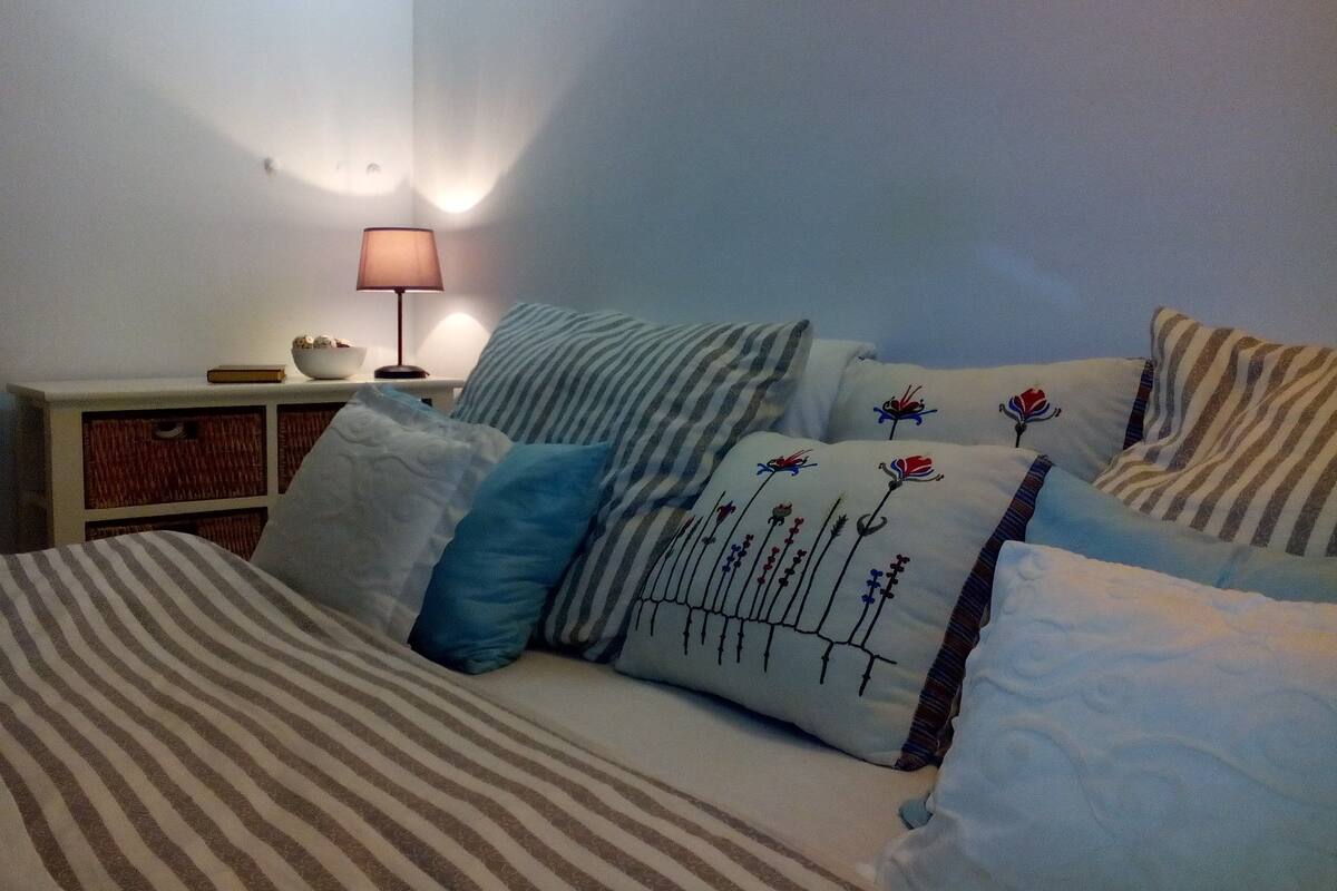 Un dormitorio con cama, almohadas, una mesita de noche con lámpara y una lámpara de pared.