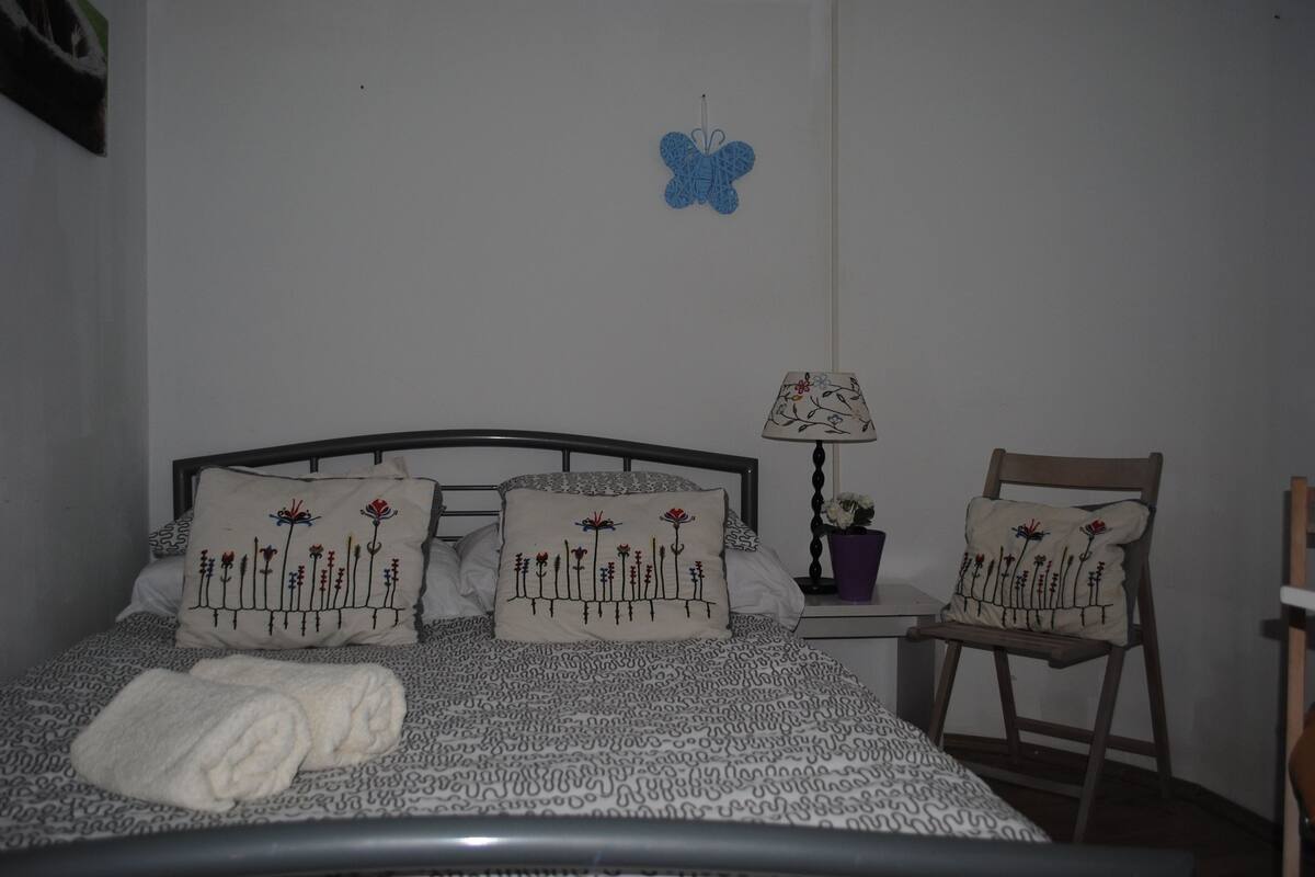 Una cama bien tendida con ropa de cama estampada, dos almohadas con diseños florales, un adorno de pared con forma de mariposa azul, una lámpara de noche y una silla con un cojín floral.