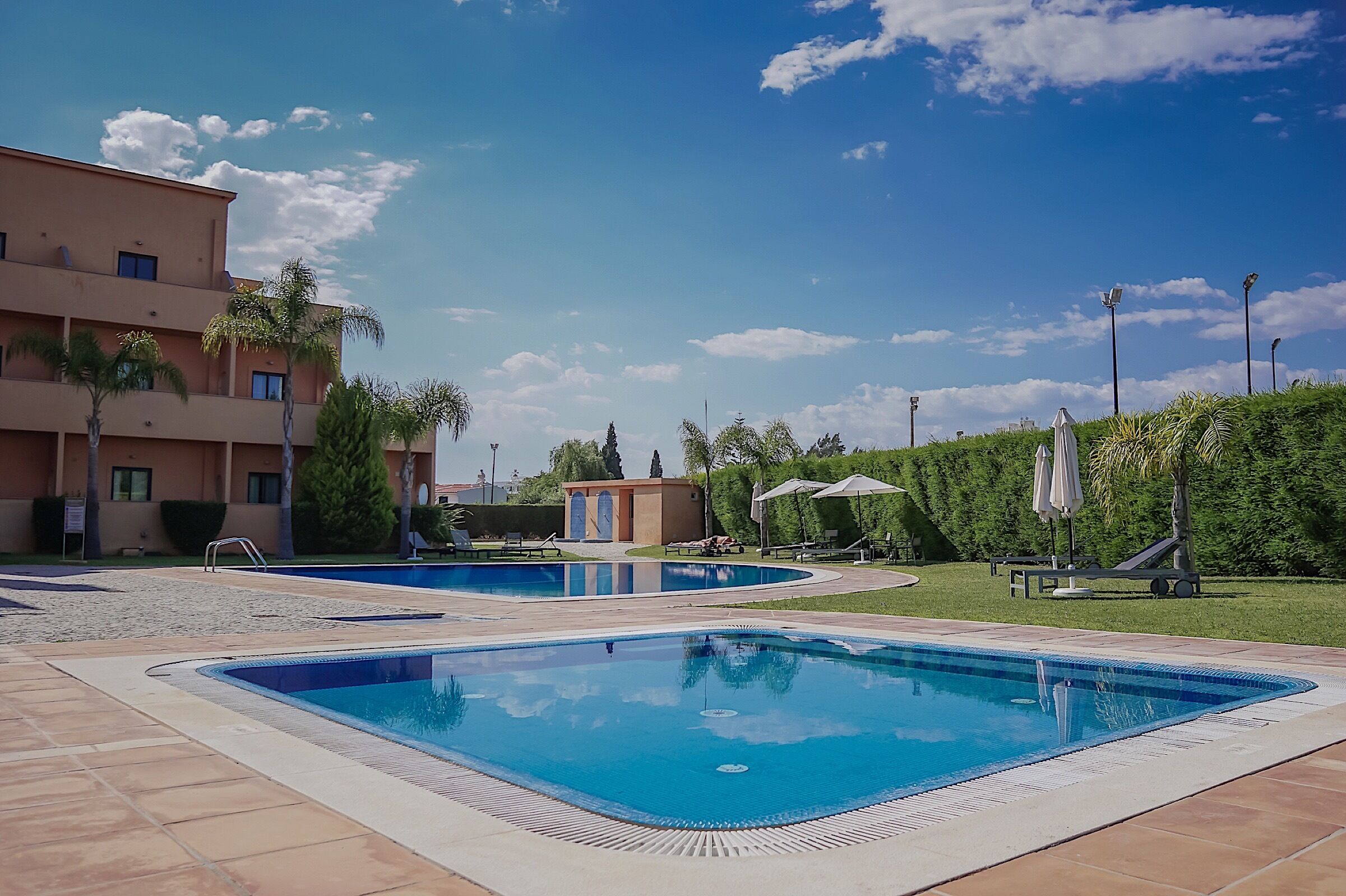 A swimming pool area with multiple pools, a building with balconies, and a well-maintained lawn with trees and umbrellas.