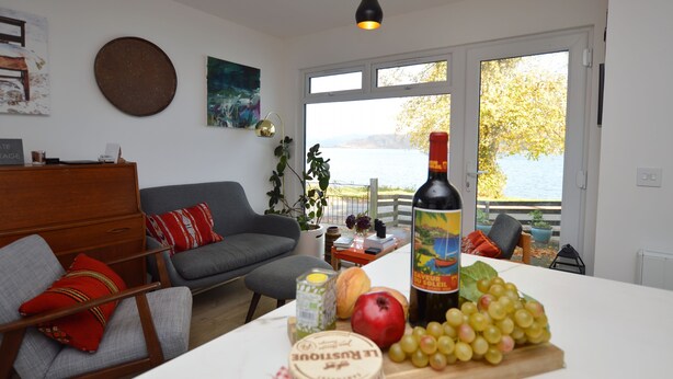 A living room with a sofa, a dining table with food, a bottle, and a view of a lake.