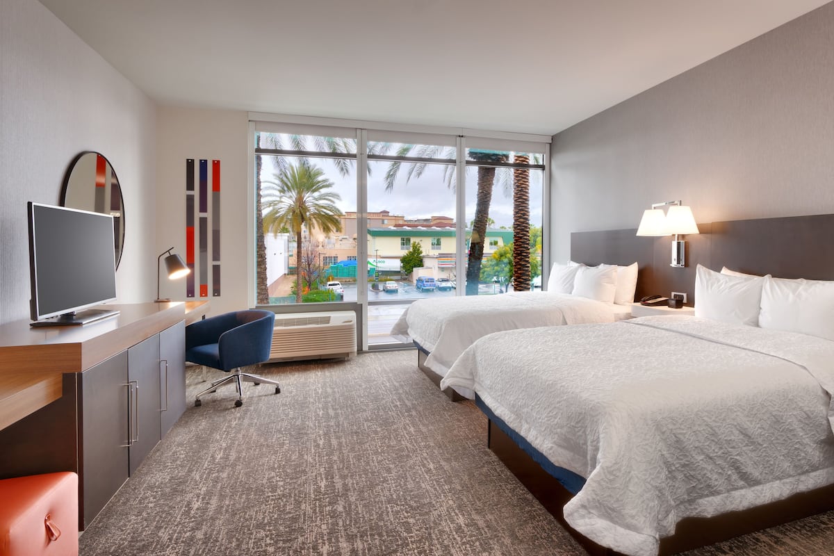 Une chambre d’hôtel avec deux lits, une télévision, un bureau, une chaise et une vue sur des palmiers et un bâtiment à l’extérieur.