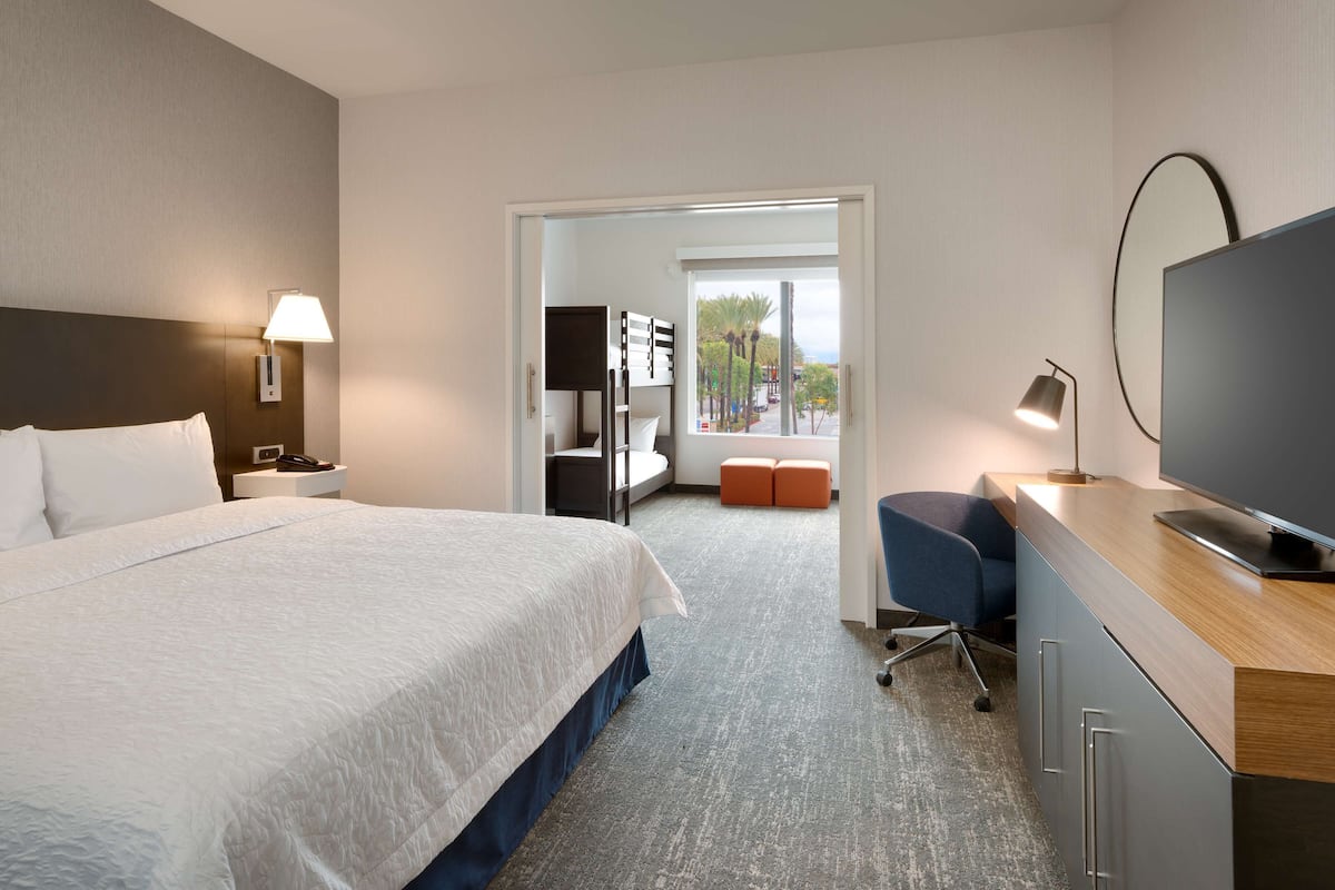 Une chambre d’hôtel avec un grand lit, un bureau avec une télévision, une chaise et une fenêtre donnant sur des palmiers.