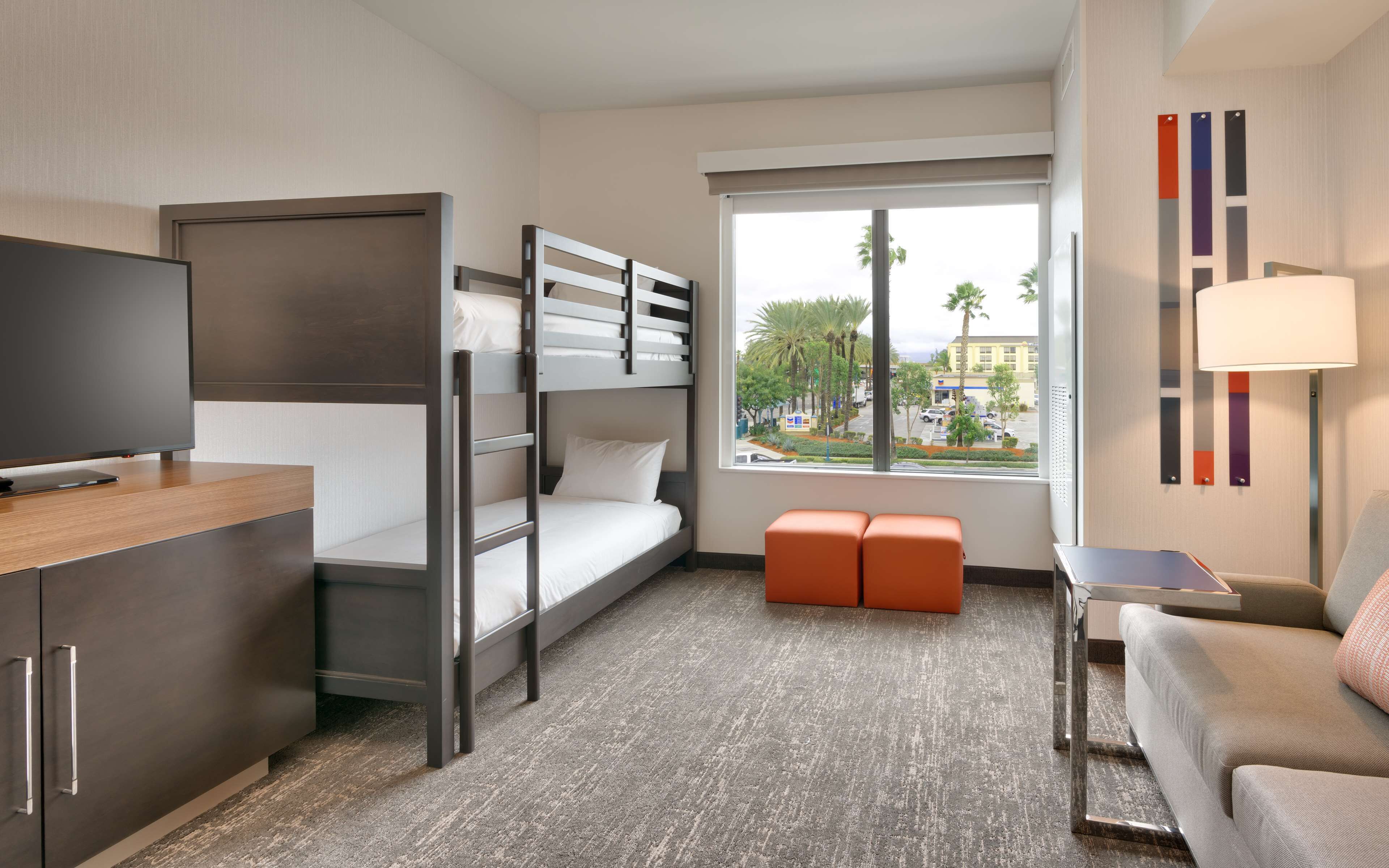 A hotel room with a bunk bed, a desk with a flat-screen TV, a sofa, a small table, and a window with a view of palm trees and buildings.