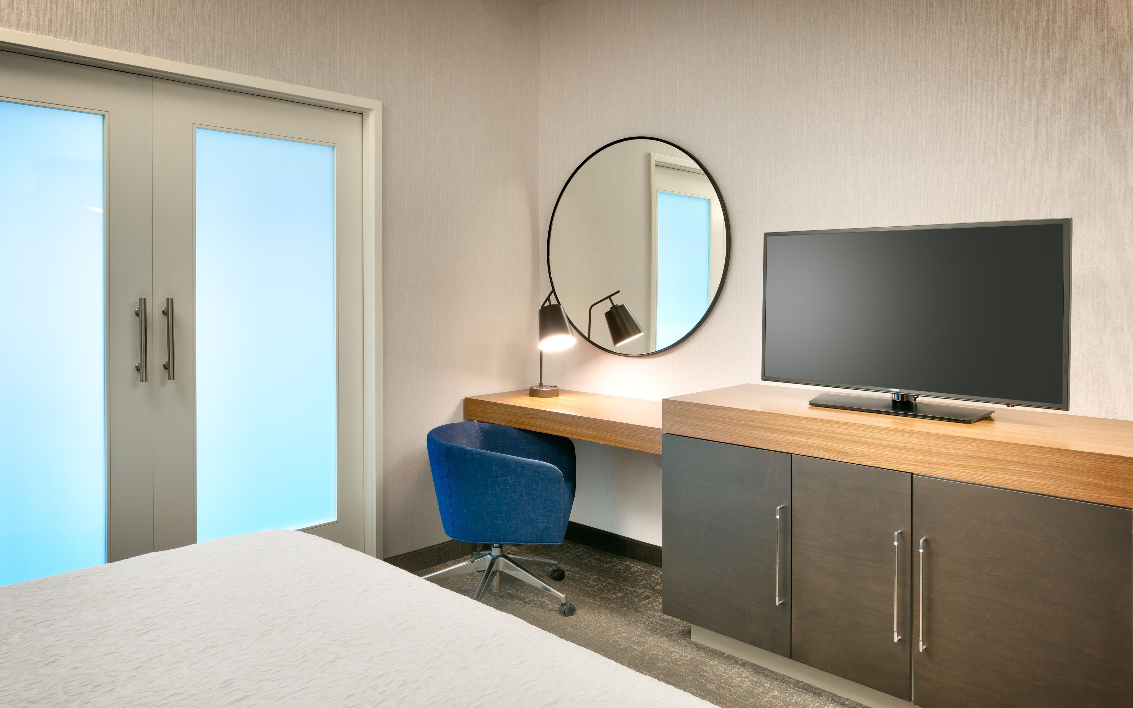 A hotel room with a bed, a desk with a TV, a chair, a desk lamp, and a mirror.