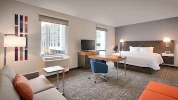Quarto de hotel com cama, escrivaninha com computador, cadeira, sofá e janela com persianas.