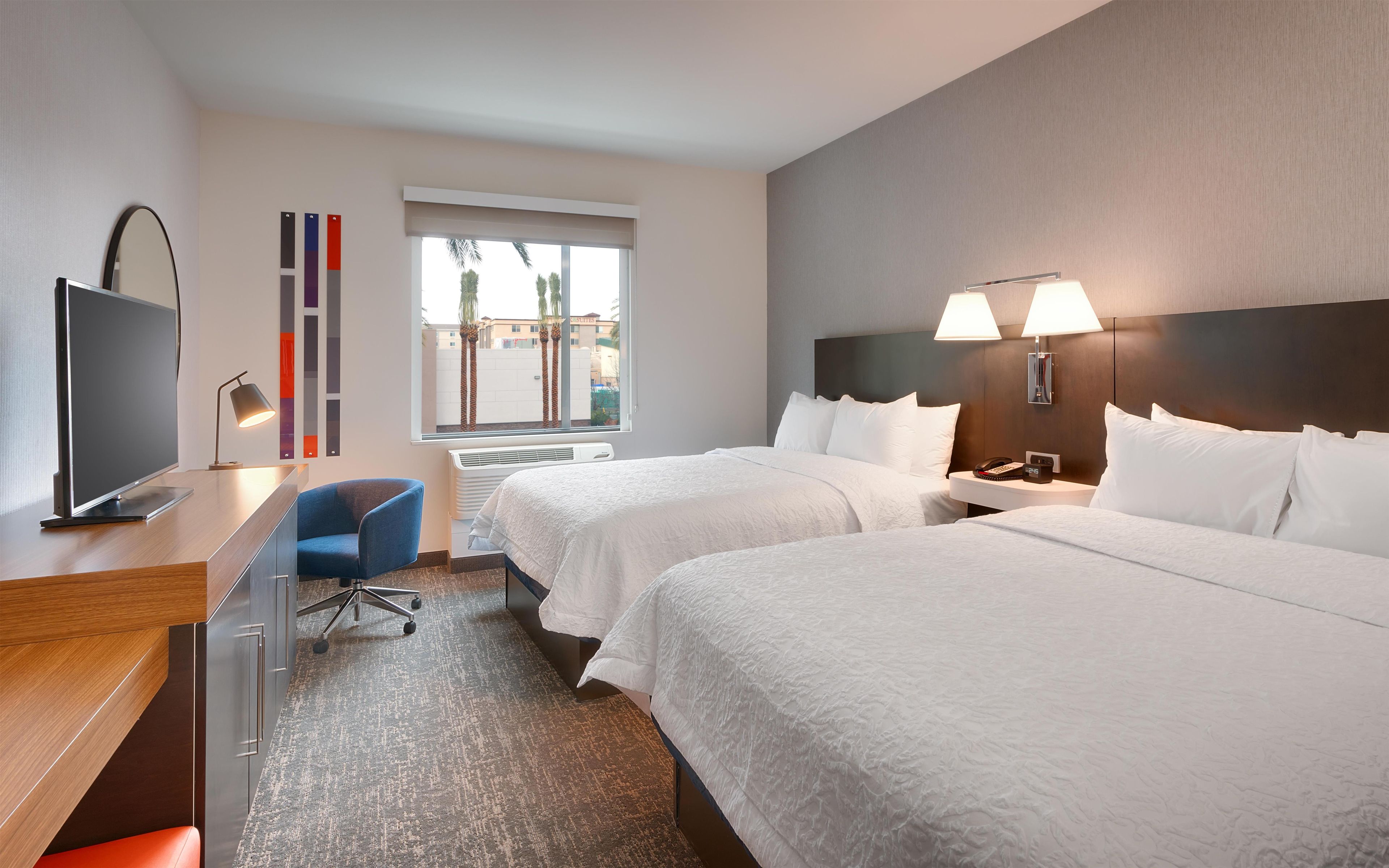 A hotel room with two beds, a desk with a flat-screen TV, a chair, and a window with blinds.