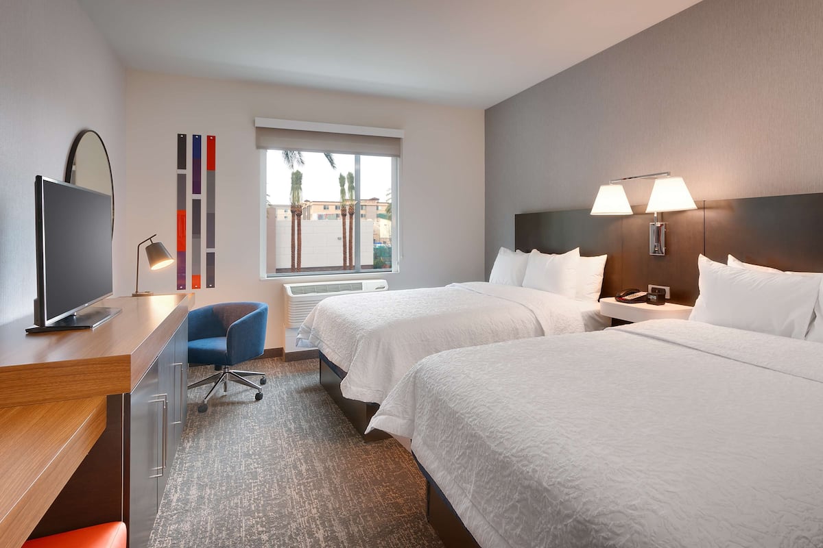 Une chambre d’hôtel avec deux lits, un bureau avec une télévision, une chaise et une fenêtre donnant sur des palmiers.