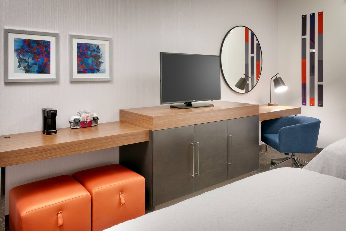 Une chambre d’hôtel moderne avec un bureau en bois, un téléviseur à écran plat, un grand miroir et deux poufs orange.