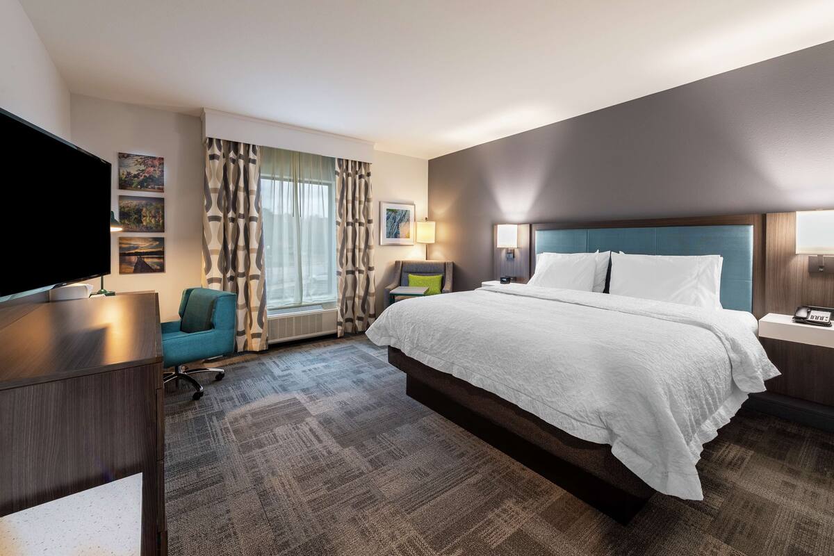 A hotel room with a large bed, a desk with a flat-screen TV, a chair, and a window with curtains.