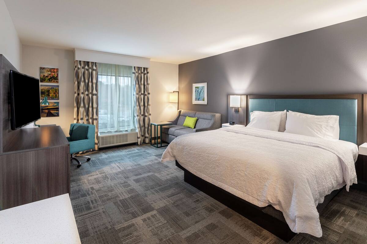 A hotel room with a large bed, a desk, a chair, a TV, and artwork on the walls.