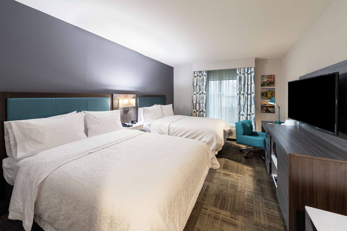 A hotel room with two beds, a desk, a television, and a window with curtains.