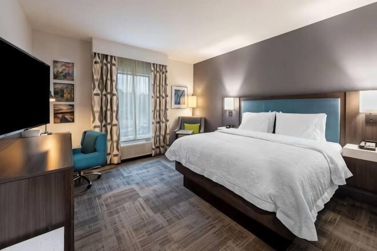 A hotel room with a bed, a TV, a desk, a chair, a window with curtains, and a nightstand.