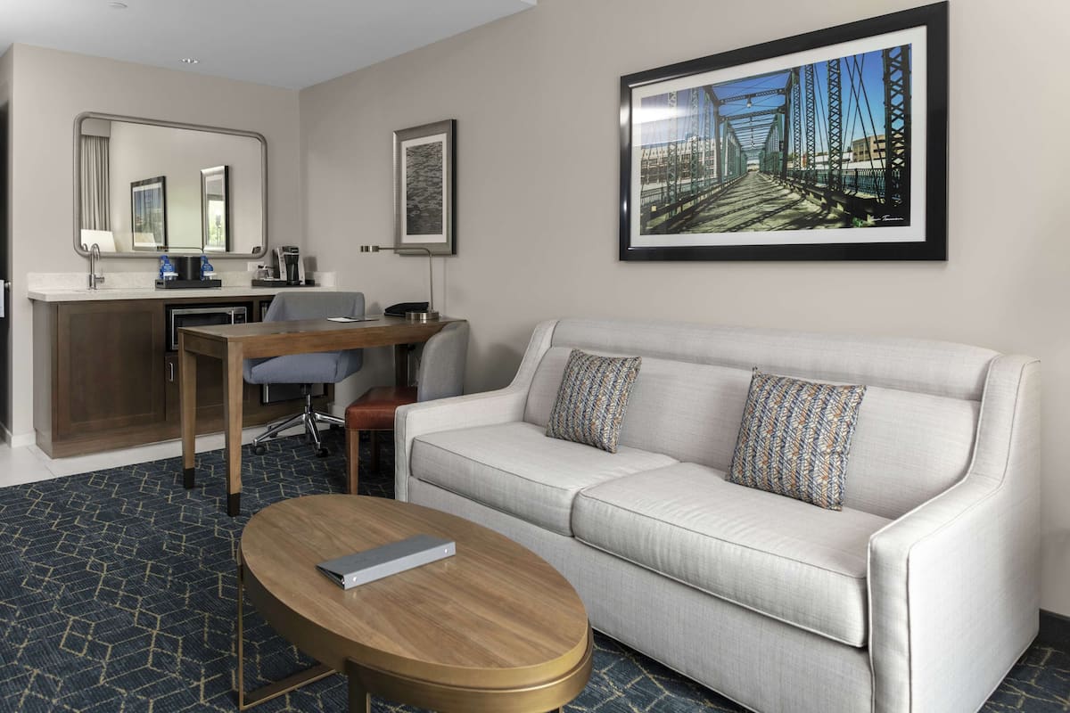 Ein Hotelzimmer mit Sofa, Couchtisch, Schreibtisch mit Stuhl, Spiegel und einem gerahmten Bild an der Wand.