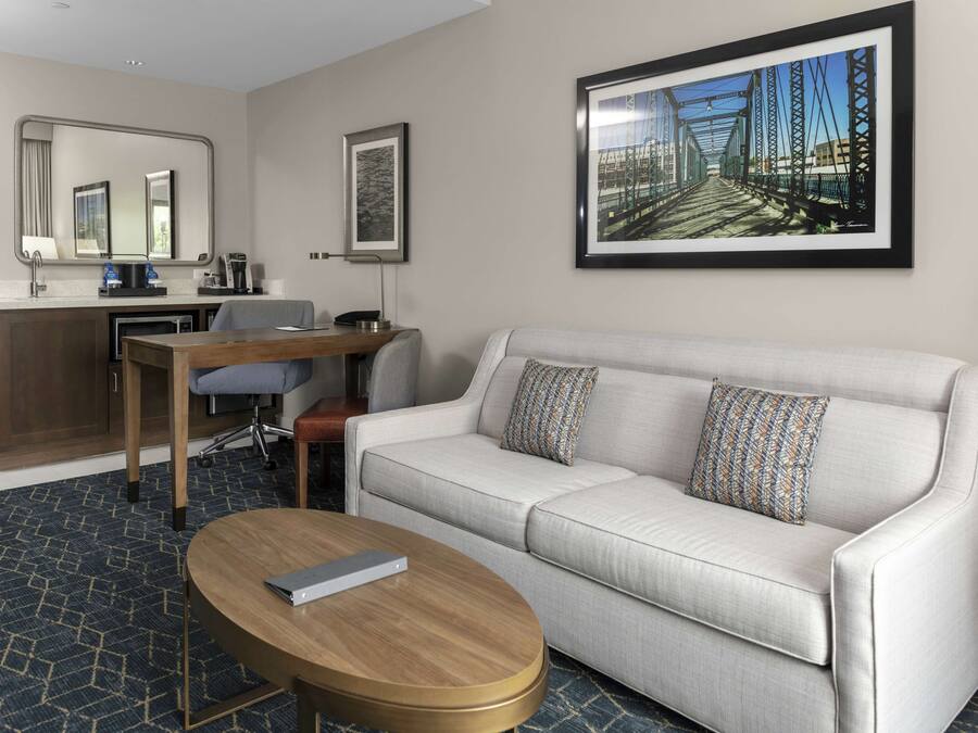 Ein Hotelzimmer mit Sofa, Couchtisch, Schreibtisch mit Stuhl, Spiegel und einem gerahmten Bild an der Wand.