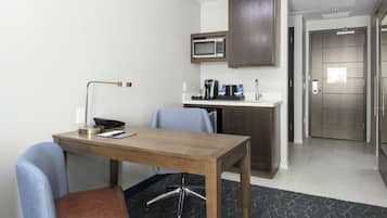 Una camera d'albergo con una scrivania in legno, una sedia blu, un forno a microonde, una macchina per il caffè e un angolo cottura.