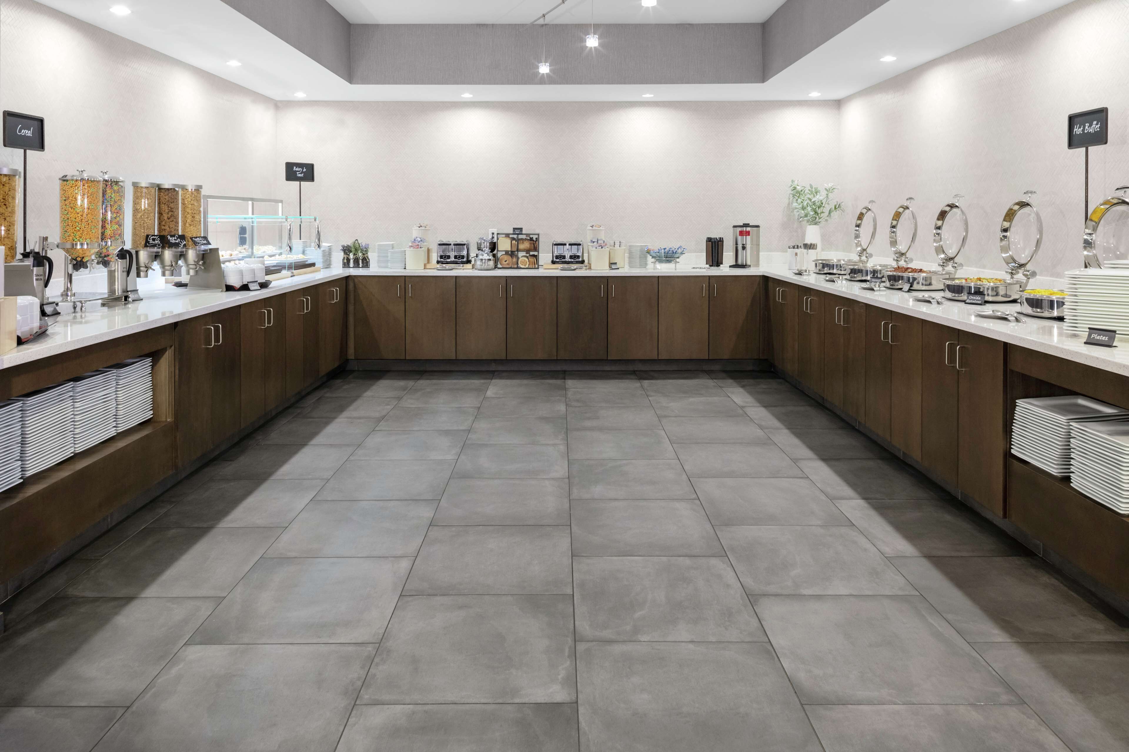 A modern breakfast area with a long counter, various food dispensers, and a clean, tiled floor.
