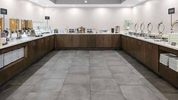 A modern breakfast area with a long counter, various food dispensers, and a clean, tiled floor.