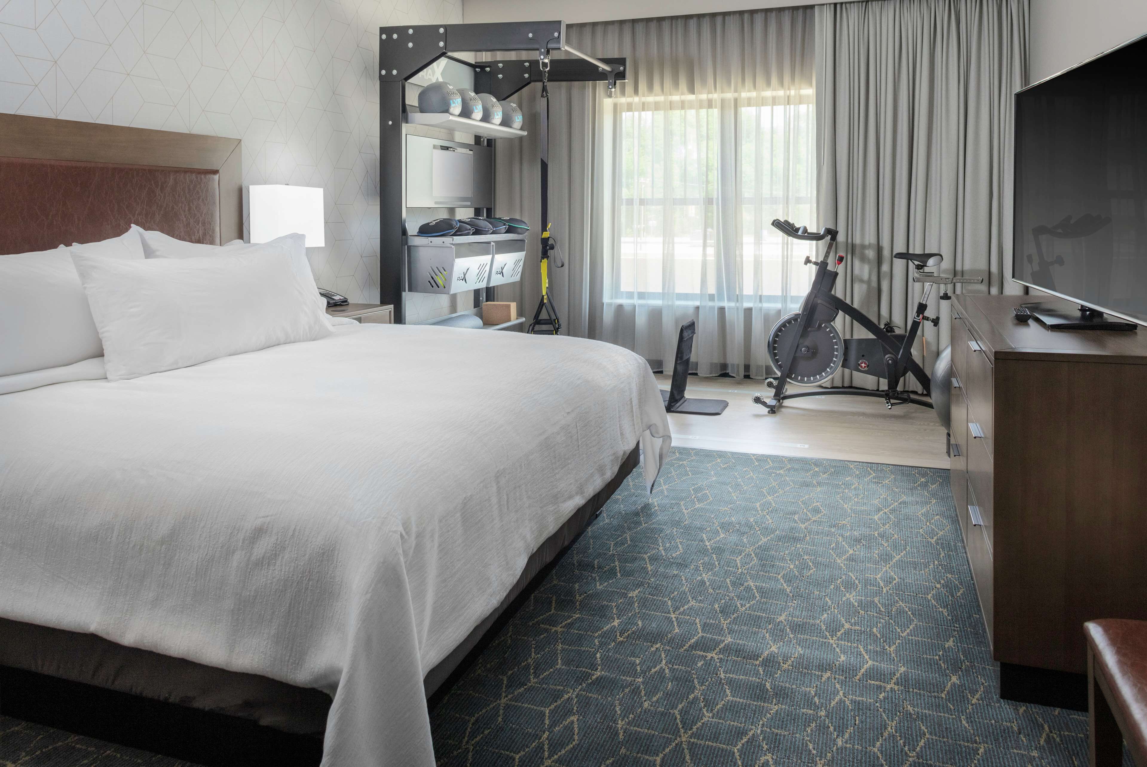 A hotel room with a large bed, exercise bike, TV, and a desk with a chair.