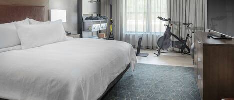 Ein Hotelzimmer mit einem großen Bett, einem Ergometer, einem Fernseher und einem Schreibtisch mit Stuhl.