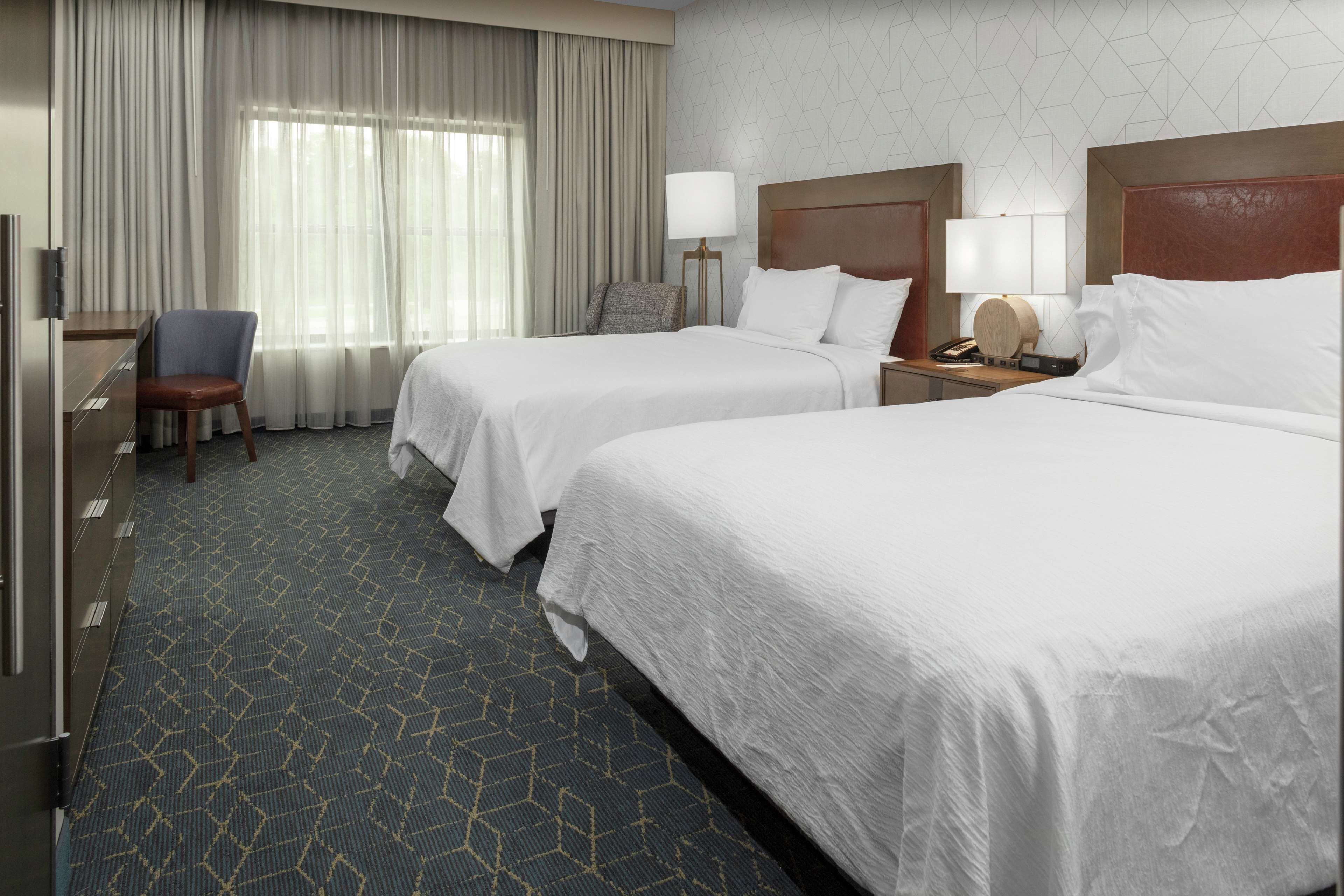A hotel room with two beds, a desk, a chair, and a window with curtains.