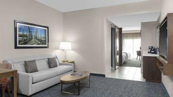 A hotel room with a sofa, coffee table, lamp, framed picture, and a view into the adjacent room.